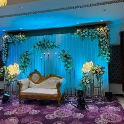A stylish stage setup featuring a white and gold diwan, floral arrangements, and dramatic blue uplighting. The large, illuminated flower props add a modern touch.