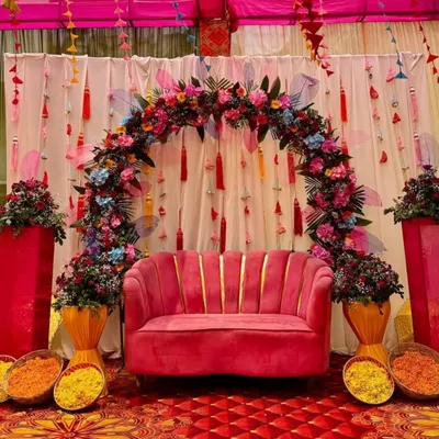 This vibrant Mehandi seating features a plush pink sofa set against a backdrop of colorful tassels and a dense floral arch. The setup includes baskets of fresh marigold petals for the rituals.