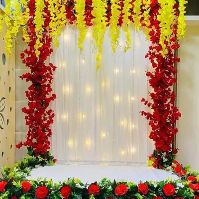 A grand arch-style backdrop made with bright red and yellow artificial flowers. The center features a sheer white curtain embedded with fairy lights, providing a luminous and festive background for your Ganpati idol.