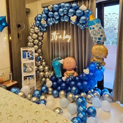 This is a full setup for an "It's a boy" baby shower. The decoration includes a blue and silver chrome balloon arch, transparent "BABY" blocks, a neon sign, and baby-themed foil balloons.