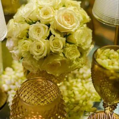 A close-up of a classic and elegant centerpiece. A tight bouquet of off-white roses is surrounded by the warm glow of candles in textured gold holders, creating a timeless and romantic look.