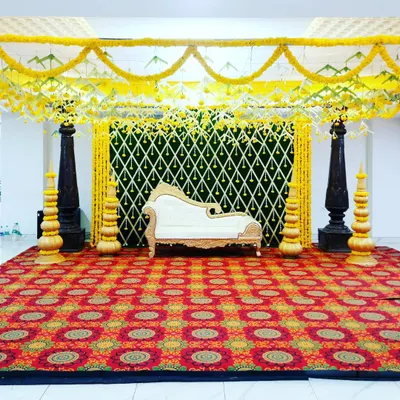 A full view of a traditional mantap setup, perfect for pujas or wedding rituals. The design is defined by its dark, carved pillars, a canopy of yellow marigolds, and a vibrant patterned carpet.