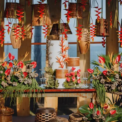 A stunning cake table from a tropical-themed engagement party. The decor features exotic red flowers, lush greenery, and hanging wicker lampshades, creating a wild yet romantic ambiance.