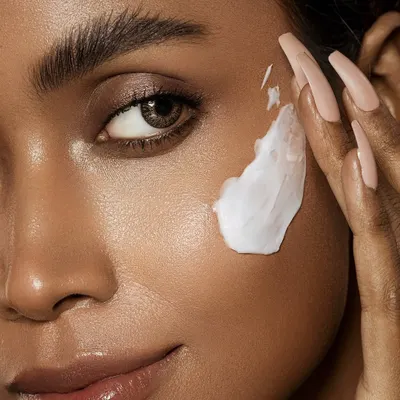 A macro shot demonstrating the application of a rich face cream. The focus is on the luxurious texture of the product against the model's radiant skin, emphasizing a moment of gentle self-care. The composition draws the eye to the interaction between the hand and face, creating an intimate feel.