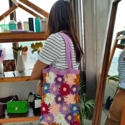 This is my multi-color daisy tote bag, shown here being worn to display its size and comfortable fit on the shoulder. The bag is covered in dozens of handmade crochet daisies in various colors, all set against a neutral background with a purple strap.