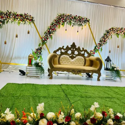 A modern and elegant stage setup for an engagement. The design features geometric arches decorated with flowers and hanging lights, with an ornate sofa as the centerpiece.