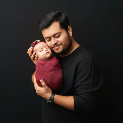 A father holds his swaddled newborn daughter close, a moment of quiet connection. I love capturing these early bonds against a simple, dark background to make the emotion shine.