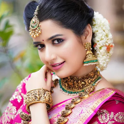 A classic South Indian bridal portrait of actress Ashika Ranganath, focusing on the rich colors of the saree and the exquisite detail of the antique gold jewelry.