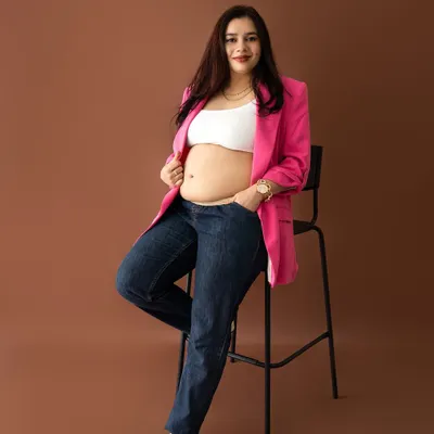 A confident and stylish maternity portrait of a mom-to-be in a pink blazer and jeans, leaning against a stool.