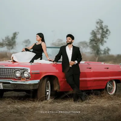 A stylish, wide-angle shot of a couple posing with a red vintage convertible in a field, capturing a classic, cinematic pre-wedding vibe.