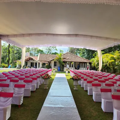 An expansive outdoor ceremony setup under a large white tent. We arranged seating for guests on either side of a central white carpet aisle, leading towards the mandap with a beautiful heritage house in the background.