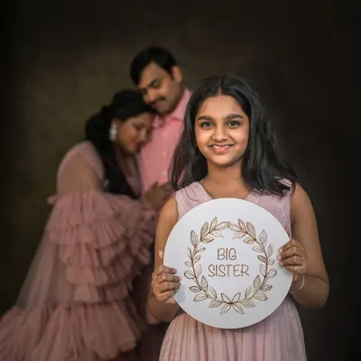 A young girl proudly holds a "Big Sister" sign, with her expecting parents softly focused in the background. This is a creative and heartwarming way to announce a new addition to the family.