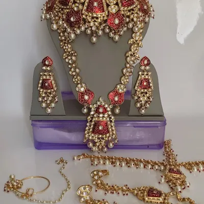 A complete bridal ensemble featuring a red meenakari choker and a longer kundan necklace. This set also includes a mathapatti, hathphool, and nath, all designed to create a cohesive and stunning bridal look.