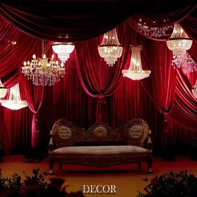A magnificent stage setup for a reception, featuring a collection of sparkling chandeliers suspended at varying heights against a wall of luxurious red velvet drapes. This design creates a focal point of pure elegance.