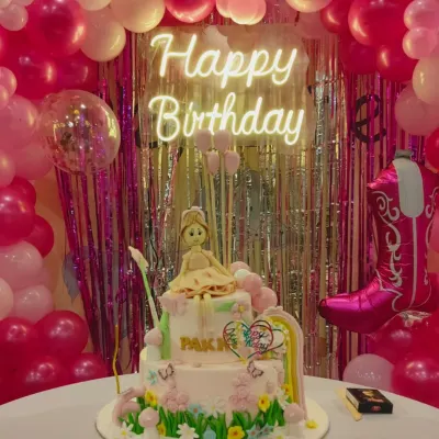 A close-up of a beautiful custom cake for a Barbie-themed party. The doll topper and intricate details are set against a backdrop of pink balloons and a neon "Happy Birthday" sign.