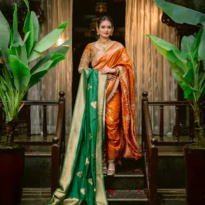 A portrait of pure elegance. The bride stands on a staircase, her vibrant orange saree and contrasting green shawl creating a stunning visual against the traditional decor.