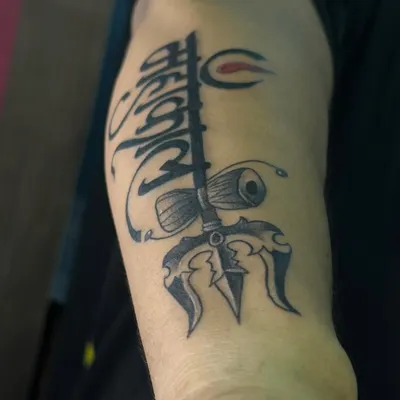 A powerful Mahakal trishul tattoo for the upper arm. This design combines the sacred trident with a damaru and calligraphic lettering, creating a classic spiritual statement.