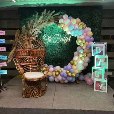 A popular baby shower decoration featuring a bohemian-style wicker chair for the mom-to-be. The setup includes a circular balloon arch in pastel colors against a green hedge wall, an 'Oh Baby!' neon sign, and 'BABY' blocks.