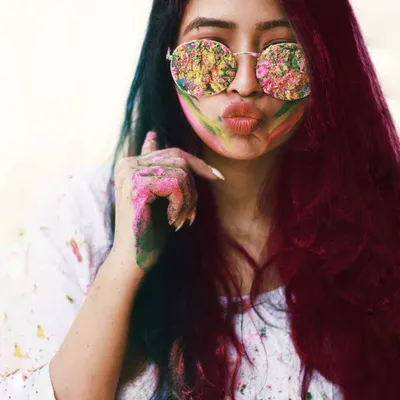 A fun and vibrant portrait celebrating the festival of Holi. The model's face and sunglasses are playfully smeared with colors, and her joyful expression captures the exuberant spirit of the celebration.