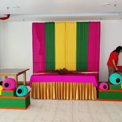 My team setting up a colorful pooja corner for a Janamashtmi celebration. The backdrop uses bright pink, green, and yellow drapes, with decorative matkis placed on either side of the main table.