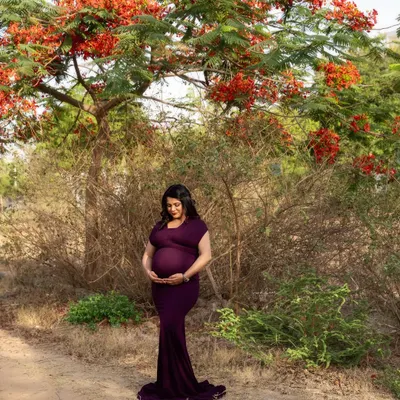 Motherhood unplugged, surrounded by nature's beauty. The vibrant red flowers of the Gulmohar tree perfectly complement the deep purple of her gown in this serene outdoor portrait.