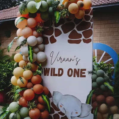 A custom "Viraan's Wild One" welcome sign with a giraffe print background. The sign is framed by an organic balloon garland in safari-inspired colors like olive green, orange, and beige, with a cute elephant cutout.
