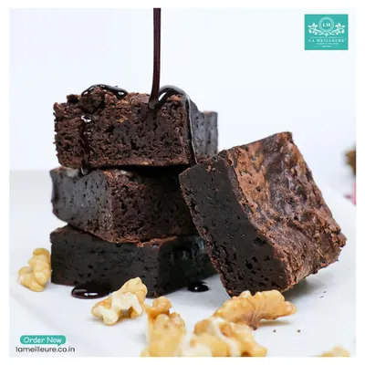 A stack of our fudgy walnut brownies with a decadent stream of chocolate sauce being poured over them. You can see the rich, dense texture in every square.