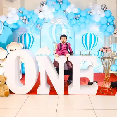 A grand first birthday celebration with a hot air balloon theme. The setup features large 'ONE' letters, a beautiful blue and white balloon arch, and adorable teddy bear accents, creating a picture-perfect scene for the birthday boy.