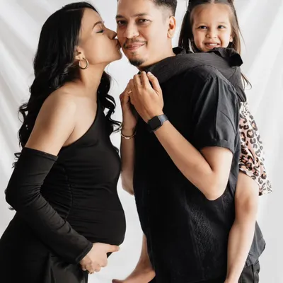 A beautiful family portrait celebrating their journey to becoming a family of four. The mother's baby bump is the focus, while the father and daughter share a fun, loving moment, all captured in a classic studio setting.