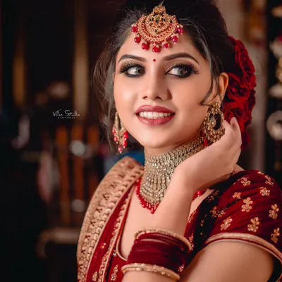 A stunning close-up of a bride in her red wedding lehenga. Her elegant pose, beautiful smile, and the intricate details of her maang tikka and choker necklace are the focus of this classic bridal portrait.