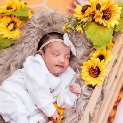 A sweet newborn baby girl in a white outfit, sleeping peacefully in a crate surrounded by sunflowers.
