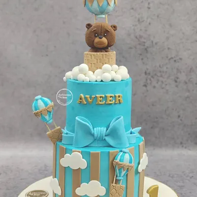 A grand two-tiered hot air balloon cake for a first birthday. This impressive non-fondant cake features a cute teddy bear in a basket, surrounded by clouds and smaller balloons.