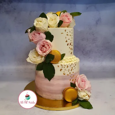 A romantic two-tier cake featuring a soft pink ombre effect on the bottom layer and a cascade of fresh pink and white roses. Gold macarons and delicate gold leaf add a touch of luxury to this wedding cake design.