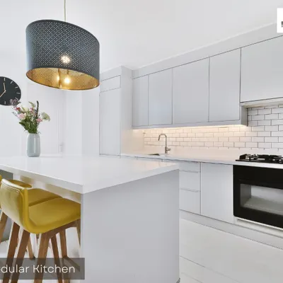 A sleek, all-white modular kitchen with an island that doubles as a breakfast bar. The design features handle-less cabinets, subway tile backsplash, and under-cabinet lighting for a bright and modern workspace.