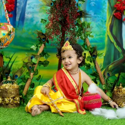 A beautiful portrait of a toddler dressed as Lord Krishna, set against a lush, painted forest backdrop. The detailed costume and golden props create a truly divine image.