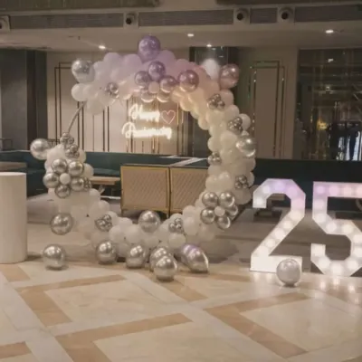 An elegant 25th-anniversary celebration with a white and silver circular balloon arch, a 'Happy Anniversary' neon sign, and large marquee numbers.