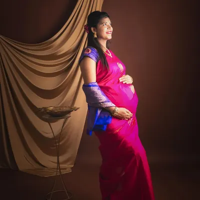 A beautiful maternity portrait of a mother-to-be in a vibrant pink and blue saree, looking down at her bump with a gentle smile.