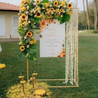 A perfect welcome for a Haldi ceremony. This entrance stand combines a sunflower and monstera leaf arch with traditional elements like hanging jasmine strings and brass lamps (diyas) on a bed of yellow petals.