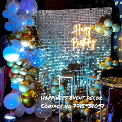A glamorous birthday setup with a silver sequin backdrop. The decor is enhanced with a balloon arrangement in blue and gold, a 'Happy Birthday' neon sign, and clear plinths for cakes or props.
