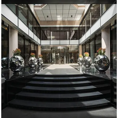 The entrance to 'The Layered Office' features a new canopy and black granite flooring leading to a revolving door. The open-air courtyard is lined with custom stainless steel planters, creating a grand and modern welcome.