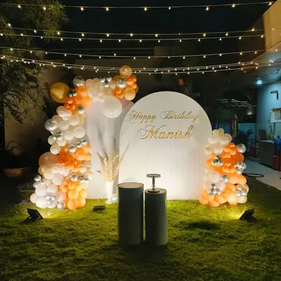 An elegant garden party setup for a birthday. I used a simple white arched backdrop, a balloon arrangement in orange, white, and silver, and overhead string lights to create a sophisticated evening look.