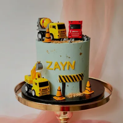 A construction-themed cake with 3D elements. This design features toy trucks and excavators on a pile of "dirt" (crushed cookies) and a road, perfect for a young vehicle enthusiast.