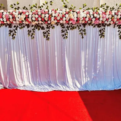 A simple and elegant stage backdrop. This setup features a white curtain with a floral and leaf border, placed on a red carpet. It serves as a clean and versatile background for any event.