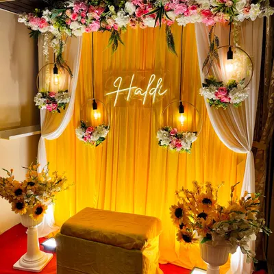 A beautiful and classic Haldi setup. This design features a bright yellow fabric backdrop, a neon "Haldi" sign, and elegant floral hoops with hanging lights.