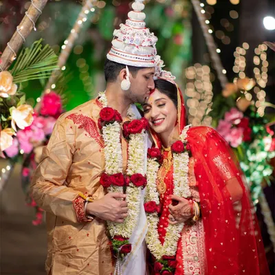 A tender moment from a Bengali wedding, with the groom kissing the bride's forehead. The traditional 'topor' and 'mukut' headwear are beautifully featured.