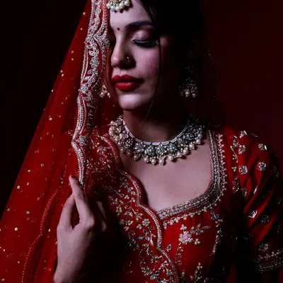 This portrait uses a deep red background and focused lighting to highlight the intricate details of the bridal lehenga and jewelry, creating a classic, elegant look.