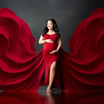 A vision in scarlet. We use flowing fabrics to add drama and movement to our maternity portraits. Here, the vibrant red gown billows around the mother-to-be, creating a powerful and artistic image against a dark studio backdrop.