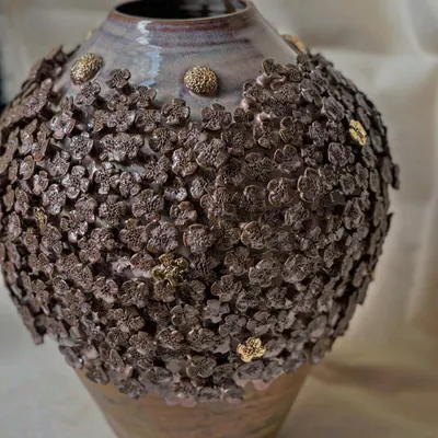 A close-up of the 'Floral Grace' vase, showing the texture and detail of the hand-applied stoneware flowers with gold accents. Each flower is attached one by one, making every vase truly unique.