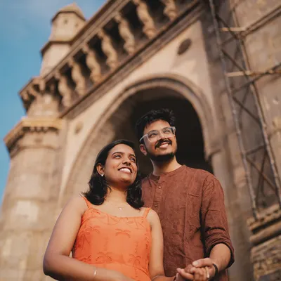 A low-angle shot of Nikit and Anusha looking up towards the sky, with the magnificent Gateway of India arching above them, capturing a moment of shared dreams in their favorite city.