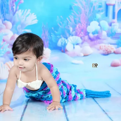 A baby girl dressed as a mermaid crawls across a dreamy, underwater-themed backdrop. The hand-knitted tail and seashell top complete this magical fantasy photoshoot.